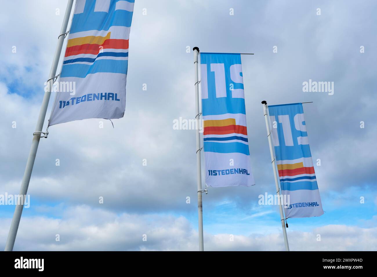 Three flags with logo in front of the elfstedenhal in `Leeuwarden against a cloudy sky. The Elfstedenhal is an indoor ice skating rink in Leeuwarden. Stock Photo